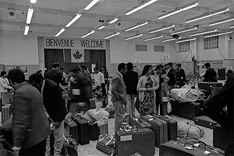 A large room crowded with People and suitcases. A Canadian flag and the word Welcome, in English and French appear on the back wall.