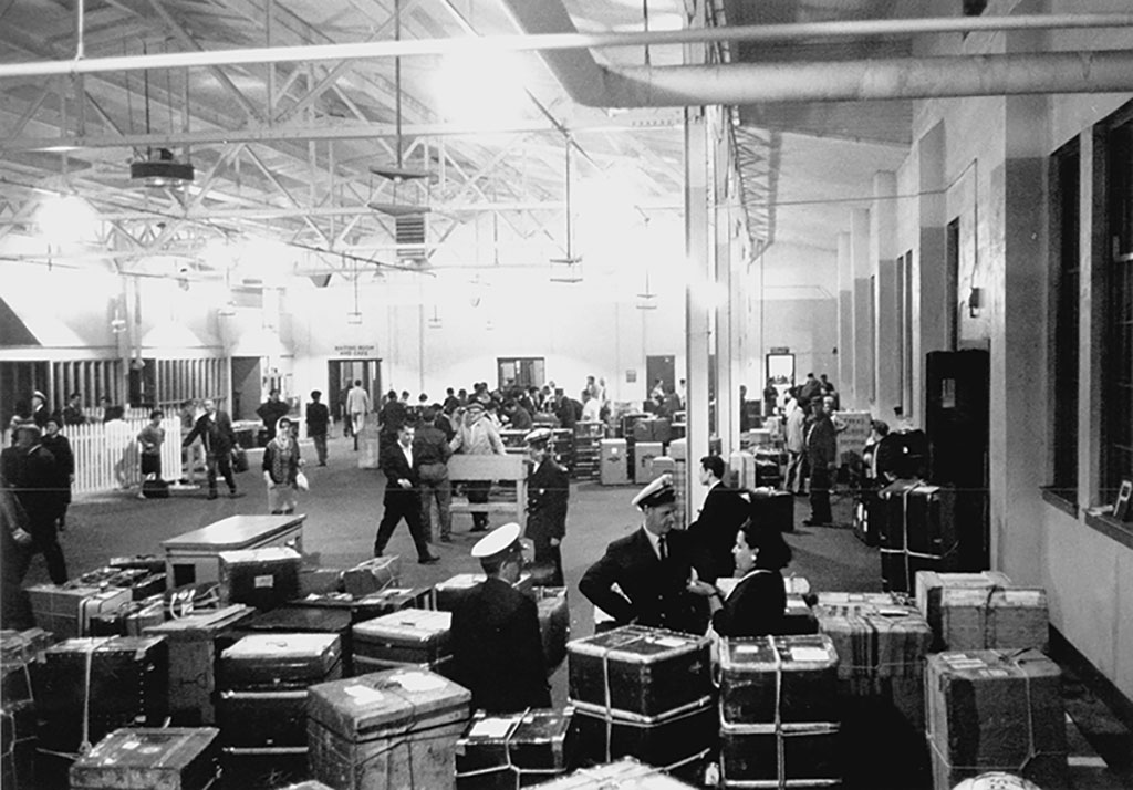 Archival image of large immigration hall filled with officers, passengers and baggage.