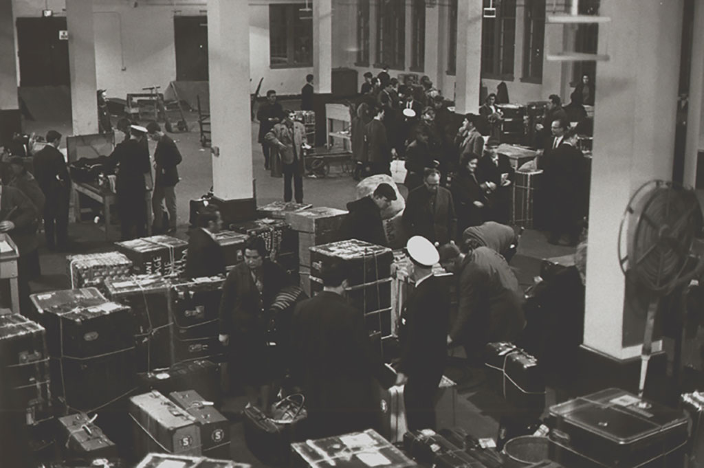 Archival image of an immigration hall, with many large travelling trunks, as viewed from above.