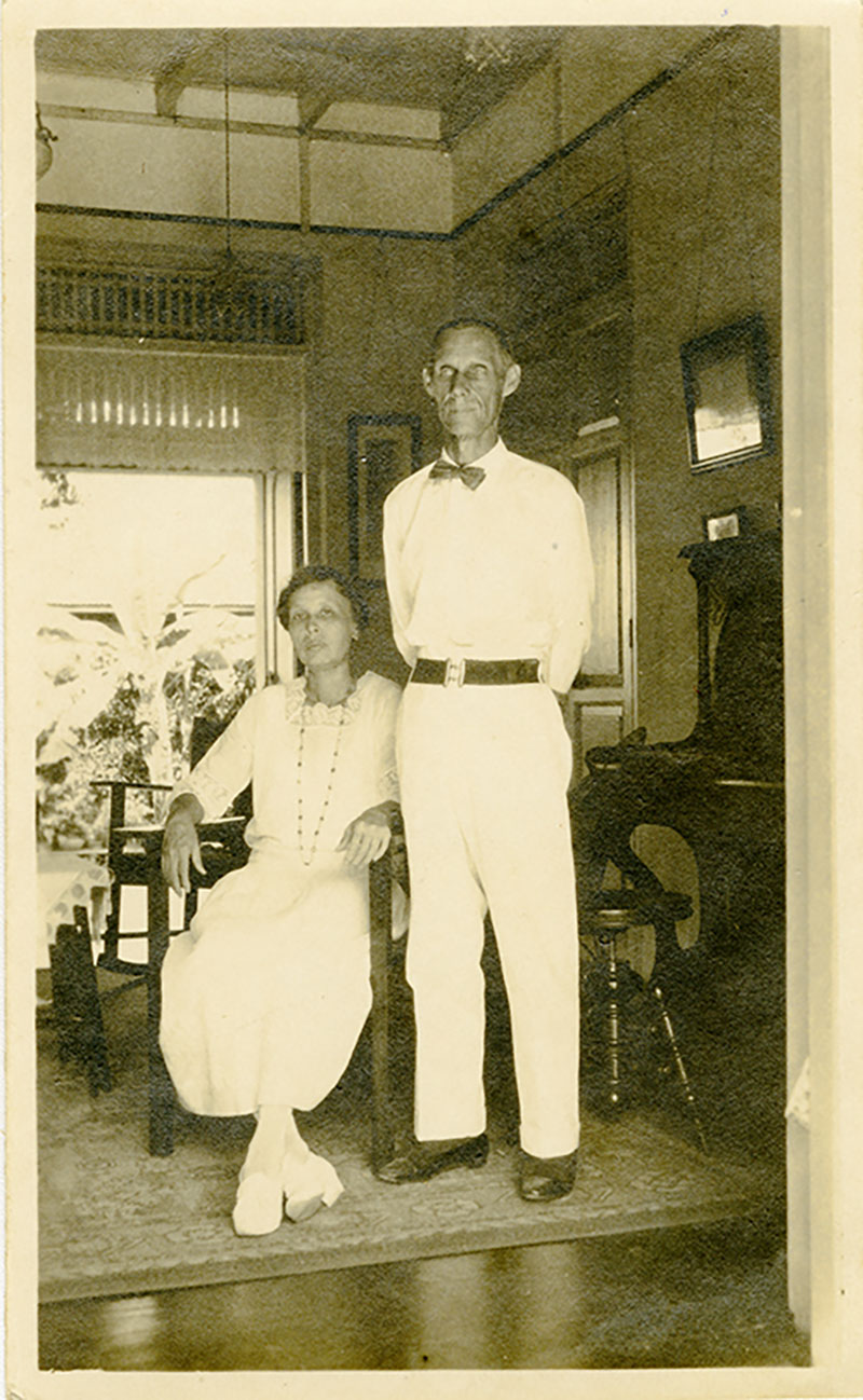A woman in white is seated, a tall man in white stands next to her.