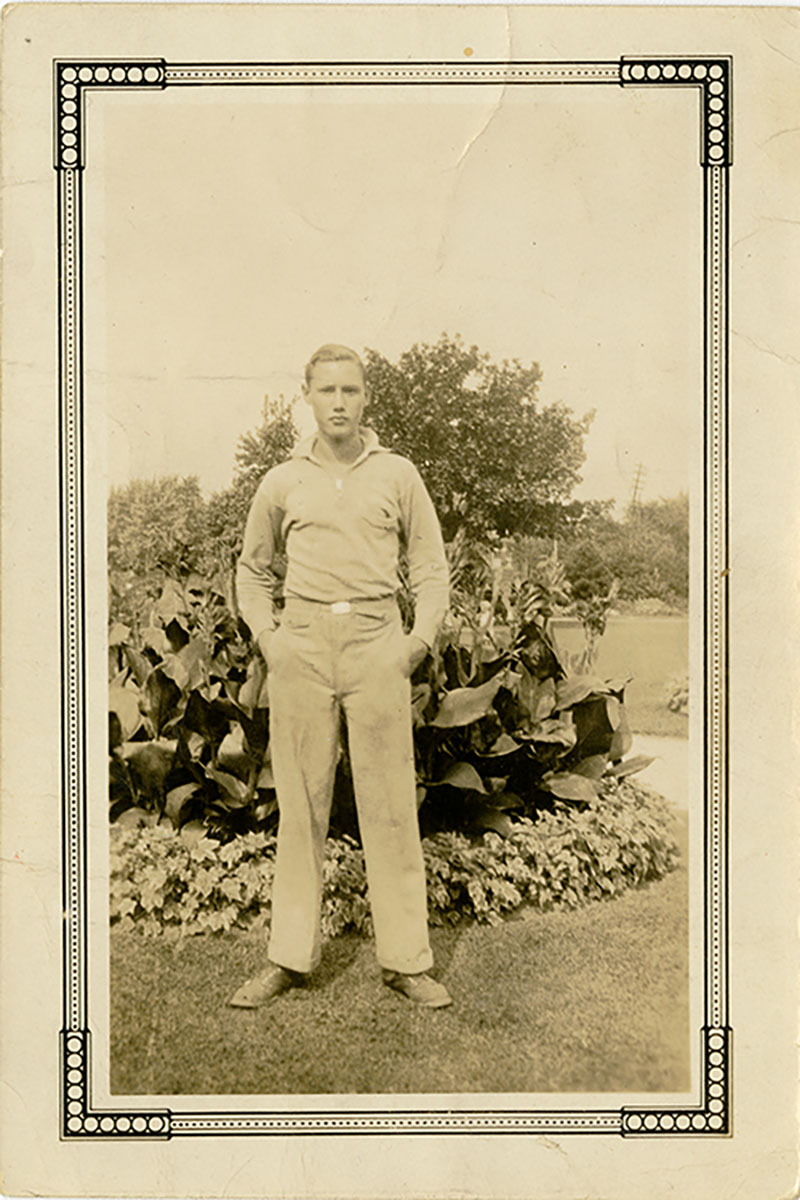 A young man with his hands in his pockets stands in front of a garden.