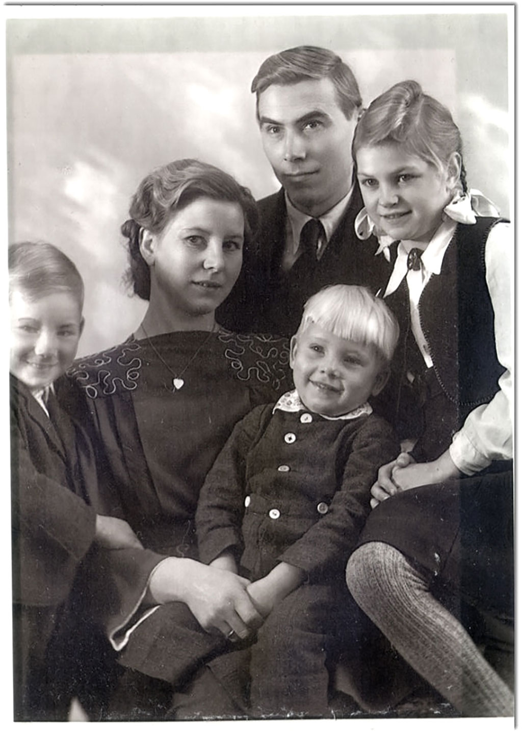 A portrait studio style black and white photograph of the Bruehler family of five with soft smiles in dress clothes.