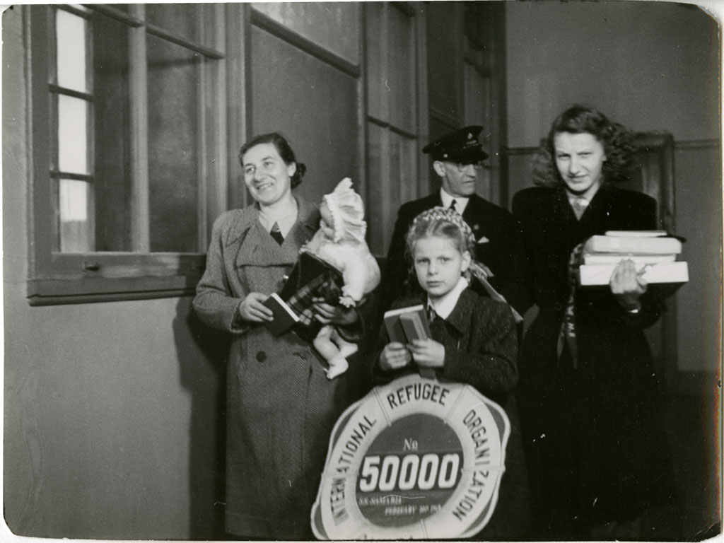 Black & white image of Latvian Displaced Person Ausma Levalds, her mother, Karline, and sister, Rasma. They are holding books and an officer stands behind them.