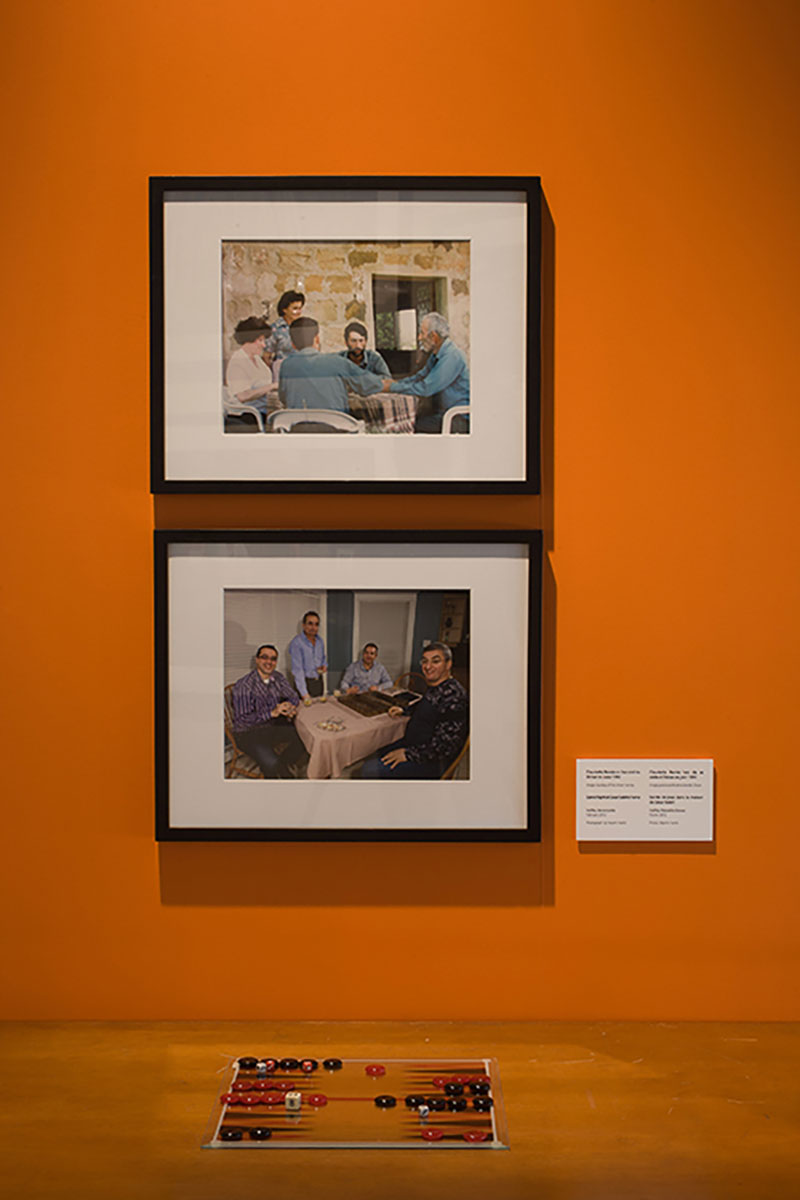 An orange-coloured wall with two framed portraits showing a group of people playing a board game. A similar board game sits on the floor in front of the portaits.