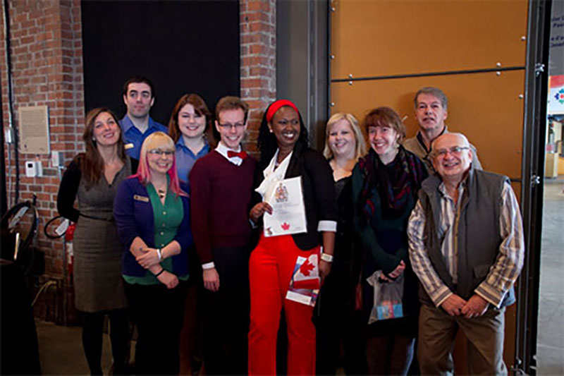 A group of smiling people surround a woman holding a Canadian flag and certificate of Canadian citizenship.