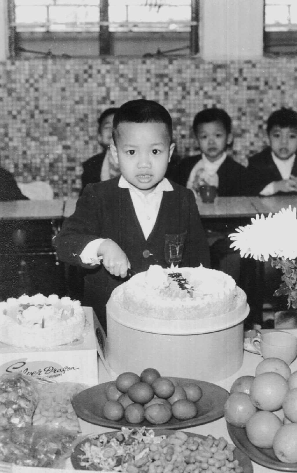 A young boy in a suit looks at the camera as he is cutting a big birthday cake.