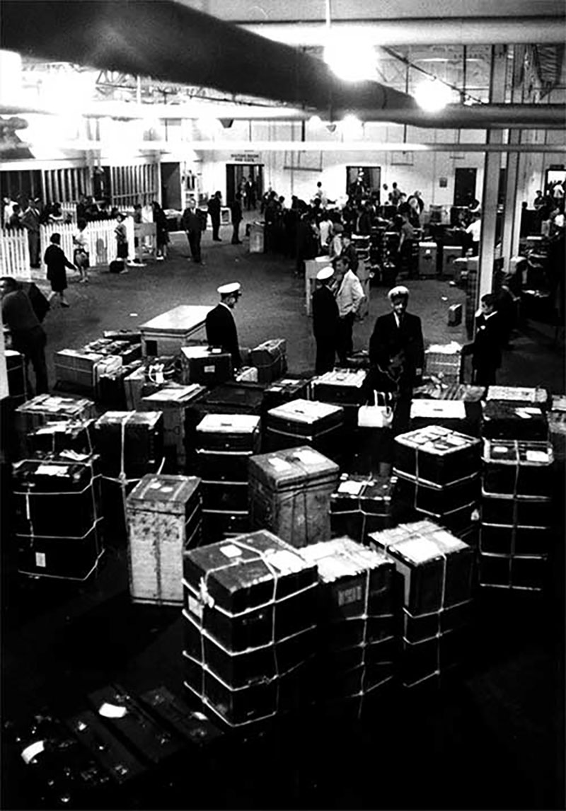 Black and white photo of large hall filled with luggage and customs officers.