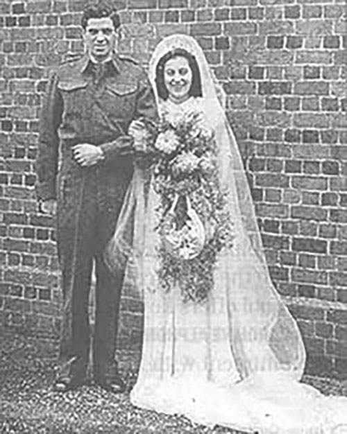 An archival image shows a bride and groom linking arms and posing for the camera, the bride has a beautiful bouquet in her other hand.