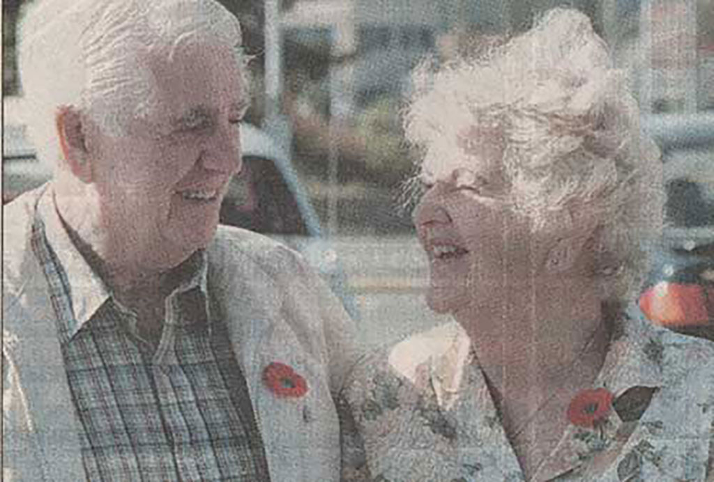 A coloured newspaper photograph shows an elderly man and woman, they are both wearing poppies and smiling at each other.
