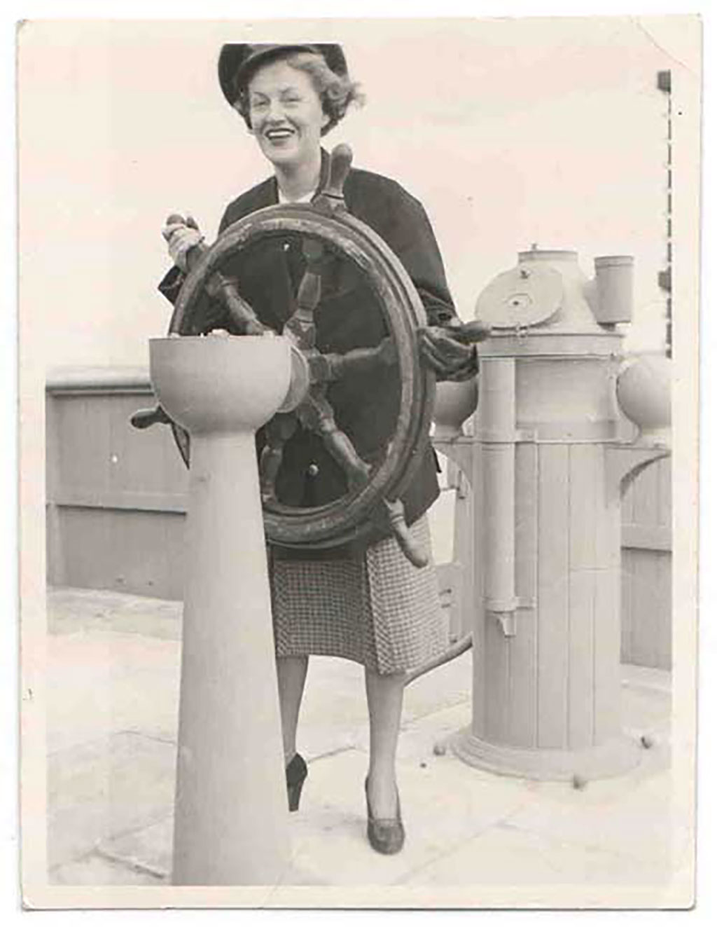 A woman in a sailor’s cap is holding on to the steering wheel of a ship, she is smiling very broadly.