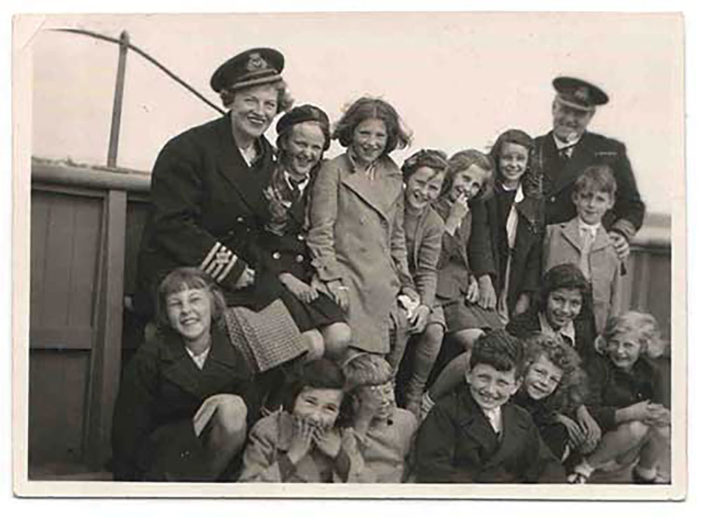 A woman in a captain’s uniform is surrounded by children of varying ages, some standing and some seated.