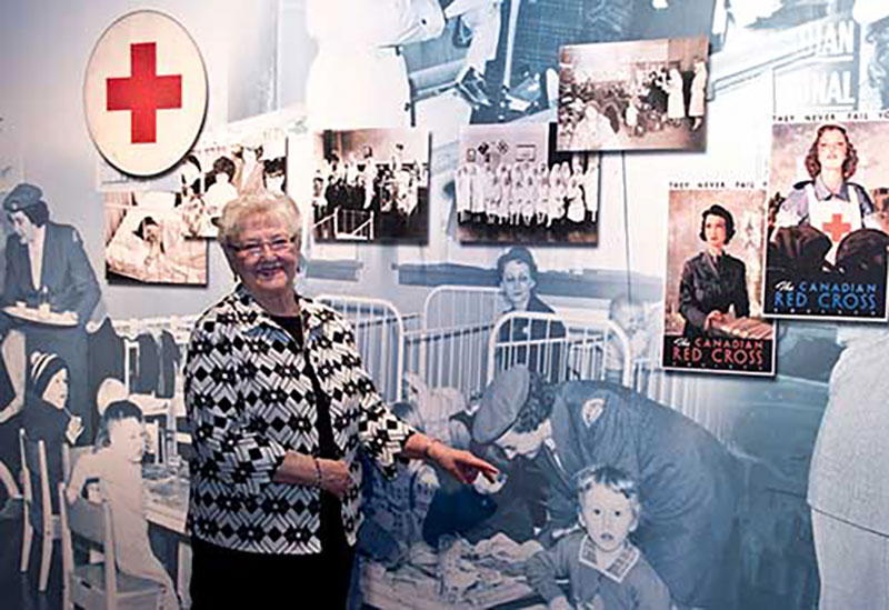 An older woman is pointing to a large mural which shows a younger version of herself as a nurse.