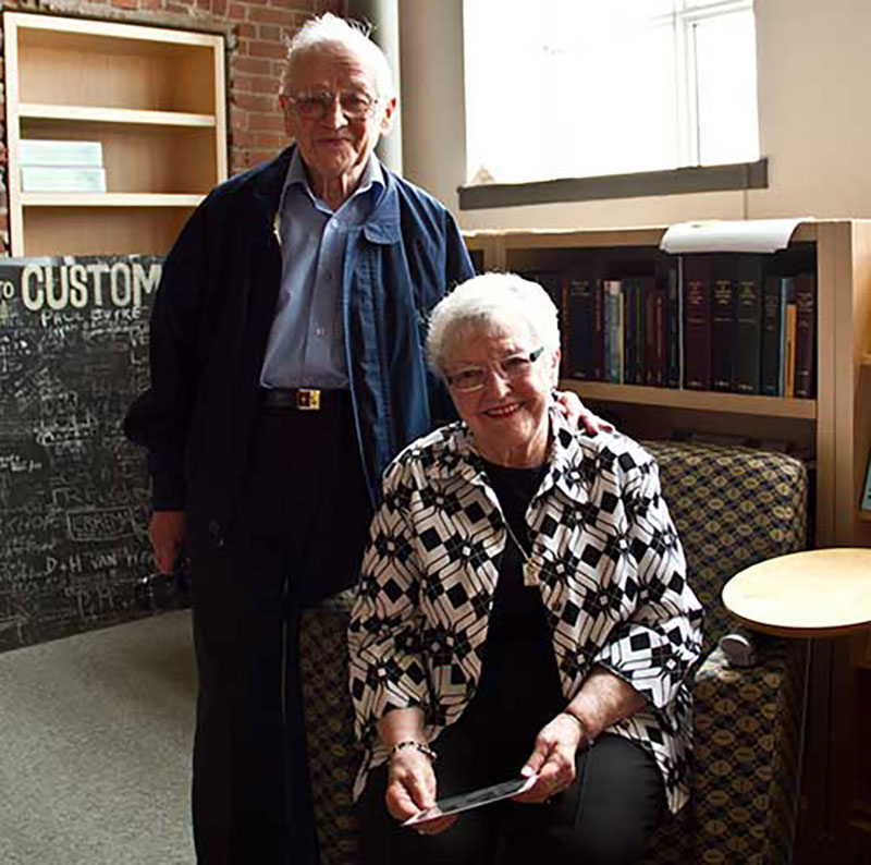 An older woman sits on a chair while an older man stands behind her with his hand on her shoulder, they are both smiling.