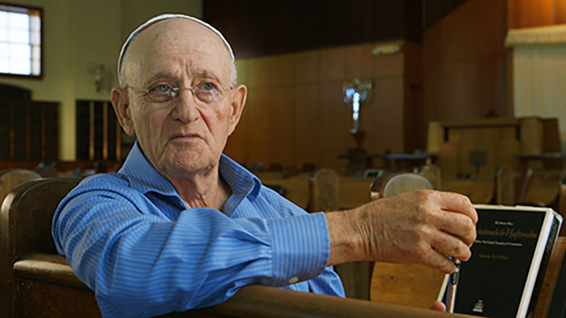 A man in a blue shirt and wearing a kippah sits in a pew of a synagogue, his body is turned towards the pew behind him.