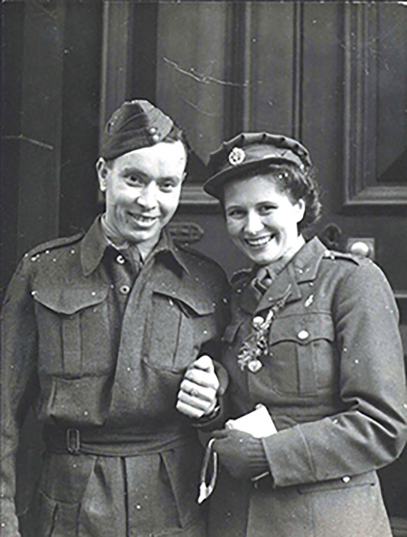 A young man and woman are standing, holding hands and smiling at the camera, both are wearing military uniforms and hats.