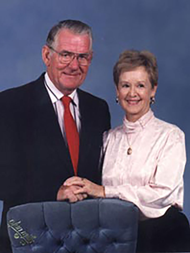 Colored portrait of a man and woman standing behind a blue chair, each with one hand resting on the chair, clasping the other. The man wears a suit with a red tie, the woman in a soft pink blouse, the background is blue.