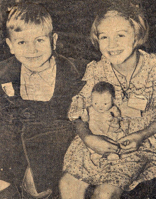 A black and white photograph of a brother and sister, the Blackmans, with big smiles. Grace holds a baby doll.