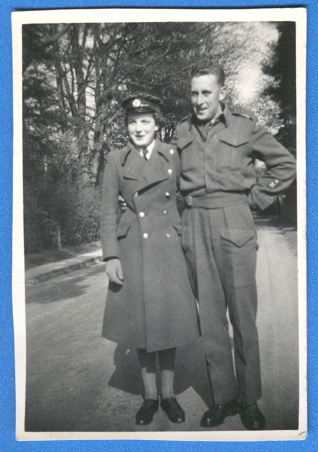 A man and woman wearing military uniform stand arm in arm on a dirt road.