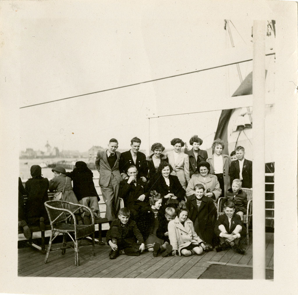 A black and white photograph of the smiling Blom family of 18 on a ship's deck. Beside them are children looking at a town.