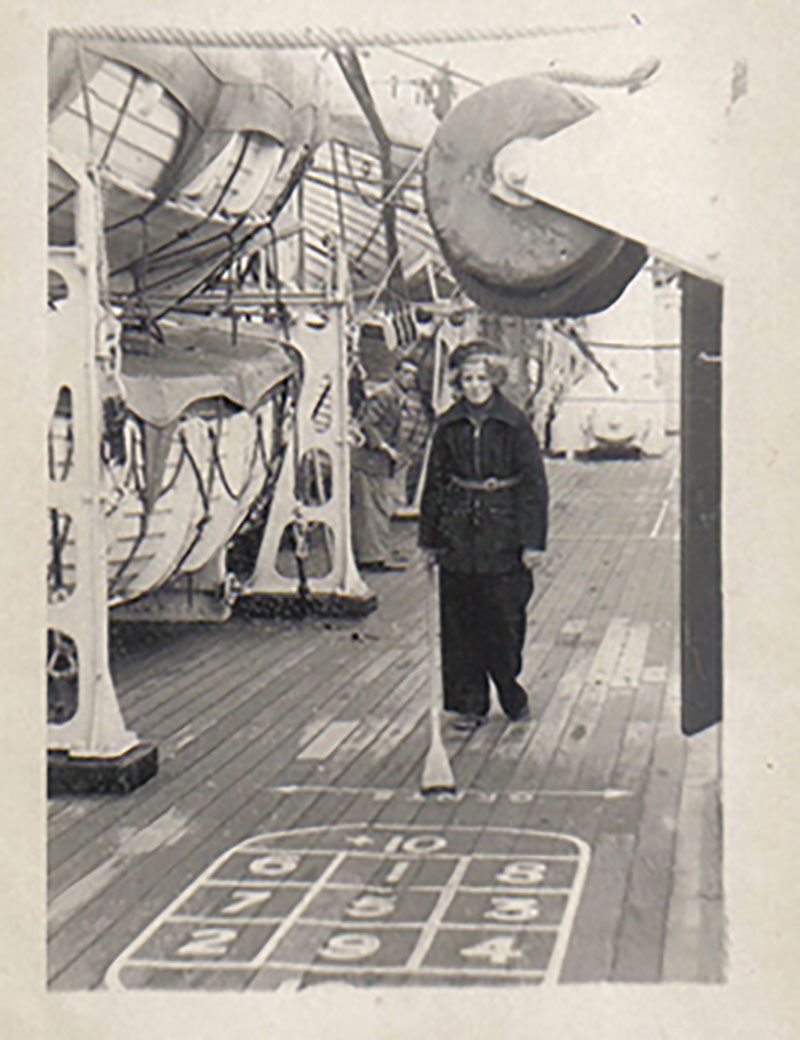 An archival photo showing a young woman in black playing shuffleboard on the deck of a ship.