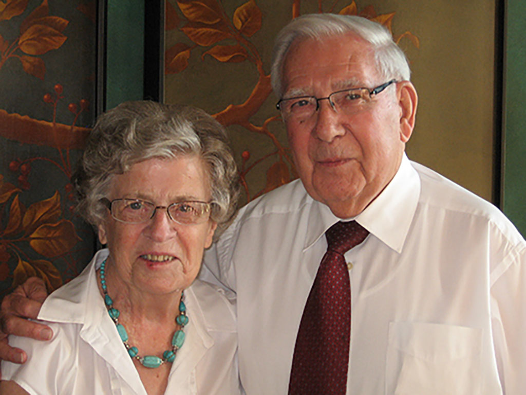 A coloured portrait of an elderly couple, they are both wearing glasses and have their arms around each other.