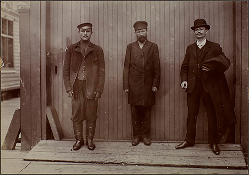 An archival image with three men standing against a wall, all wearing clothing of that era.