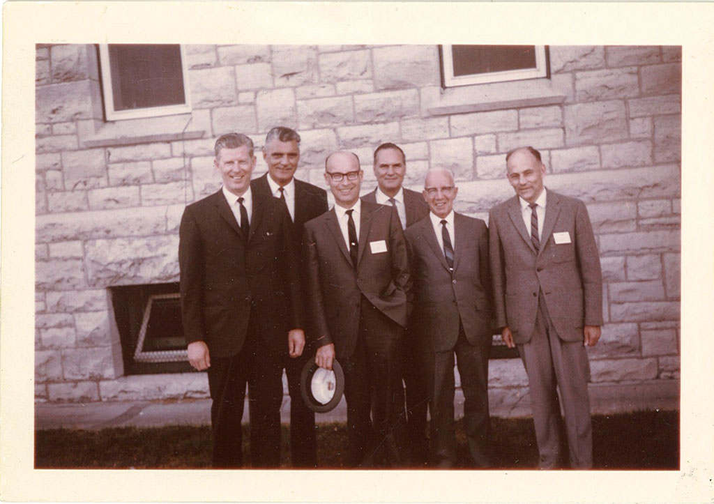 A group of men dressed in suits and ties stand in front of a stone building as they smile for the camera.