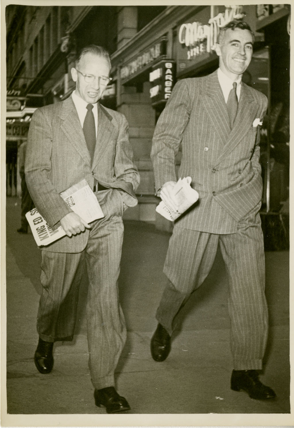 Two men walk with purpose, both holding newspapers in one hand and have the other hand in their pockets.