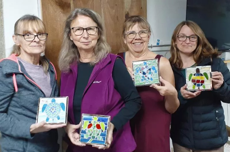 Four women participants of a glass art workshop display their handmade stained glass mosaic sea turtle panels.