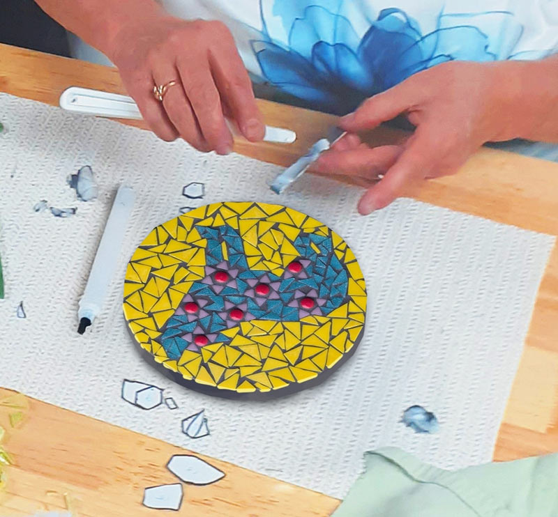 A participant carefully places coloured glass tiles onto a circular mosaic panel featuring a teal wolf design on a yellow background.