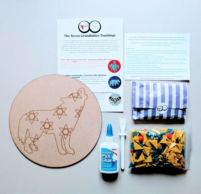 A mosaic kit laid out on a table including a circular wooden wolf template with intricate designs, a bag of coloured glass tiles, glue, a spreading tool, instruction cards, and a Seven Grandfather Teachings reference sheet.