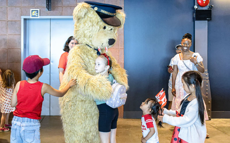 A group of children gathering around Fenton, the Museum’s teddy bear mascot, with one child hugging Fenton.