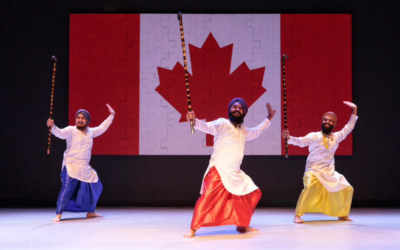 Three men dance in front of a large Canadian flag with a puzzle piece design.