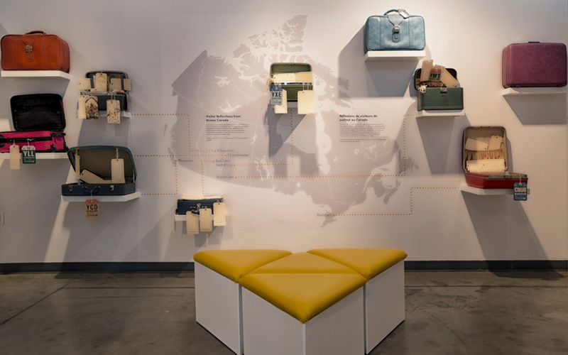 Ten pieces of luggage are displayed on a wall with text and a map of Canada.