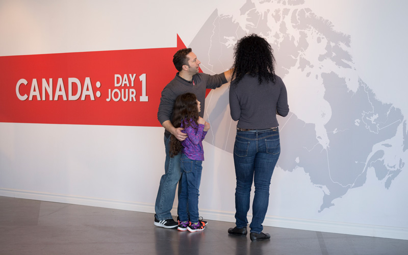 Two adults and one child look at a large map of Canada on the wall. A red sign with the exhibition title is behind them.