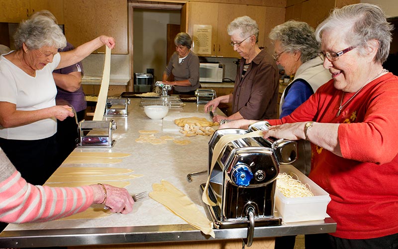 Seven women make pasta in a kitchen.