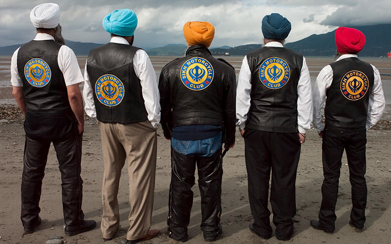 Six men stand with their backs to the camera, displaying vests that say “Sikh Motorcycle Club.”