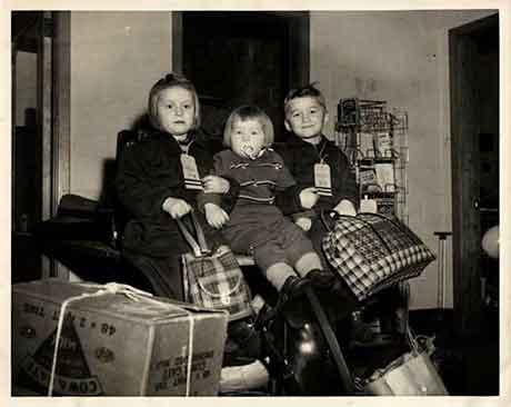 Old black & white image of three kids sitting with luggage bags, they are holding each other and look very happy.