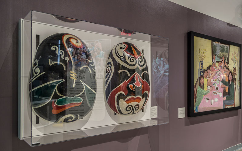 Two large masks in a glass case beside a painting of a family at a dinner table, both hung on the wall.