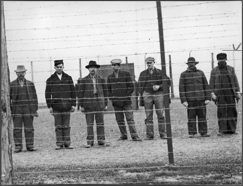 Archival image of several men standing behind a barbed wire fence.