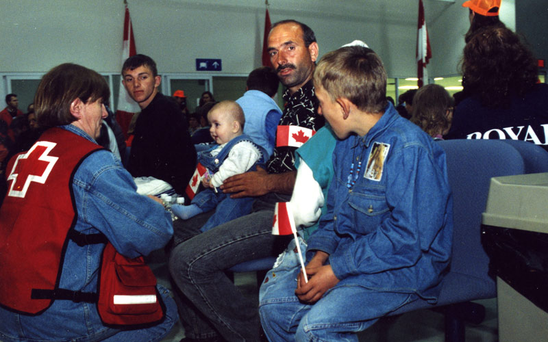 A white woman in a Red Cross vest kneels in front of a Kosovar refugee family, including a man, two teenagers, and a baby.