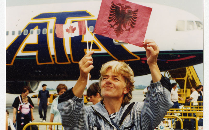 A Kosovar refugee smiling and holding up a small Albanian flag in front of an airplane.