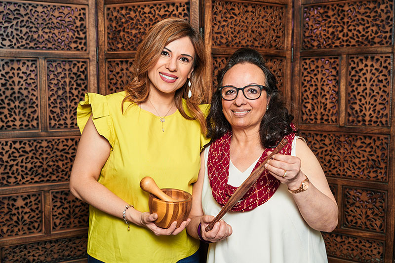 A portrait of two smiling Indian woman, one holding a mortar and pestle.