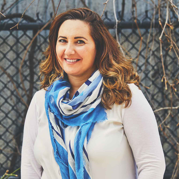 A portrait of a smiling Syrian woman with shoulder length brown hair, wearing a white shirt and blue scarf.