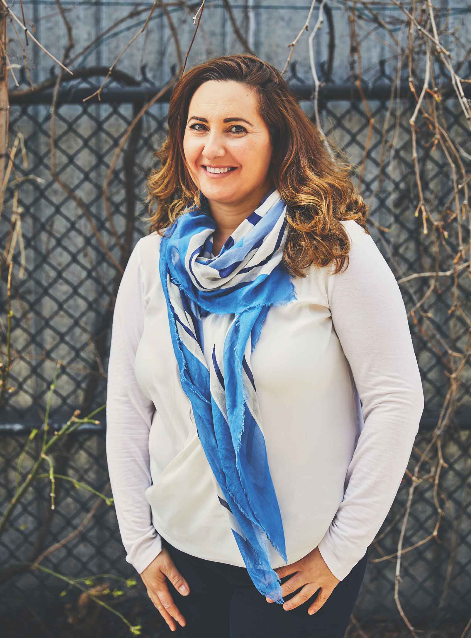 A portrait of a smiling Syrian woman with shoulder length brown hair, wearing a white shirt and blue scarf.