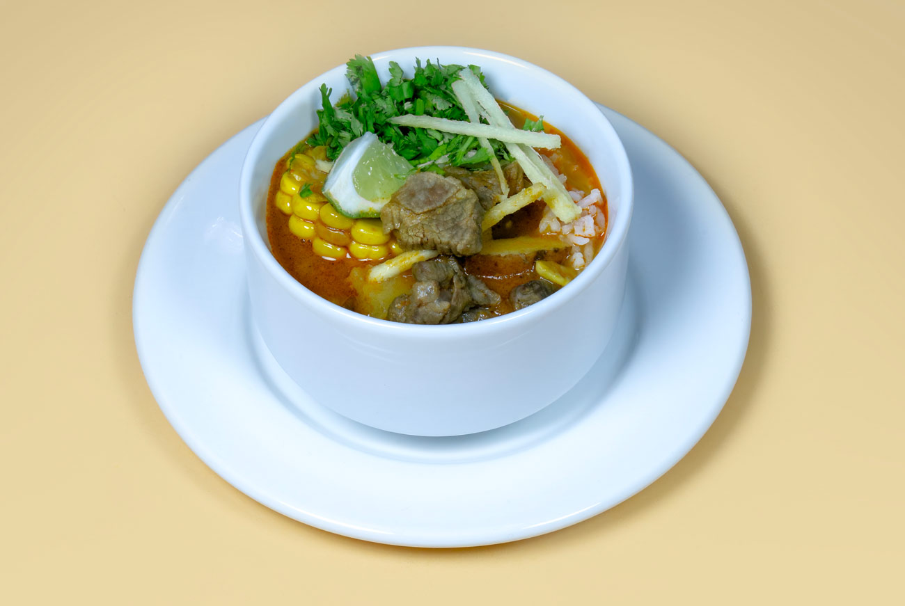 A bowl of aloo birria with reddish broth, corn on the cob, beef pieces, rice, cilantro, tortilla strips and a lime wedge.