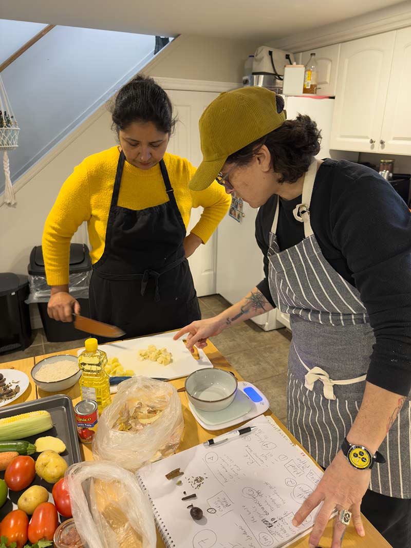 Elvira and Hina prepare food together in a kitchen, standing at a counter with ingredients and utensils.