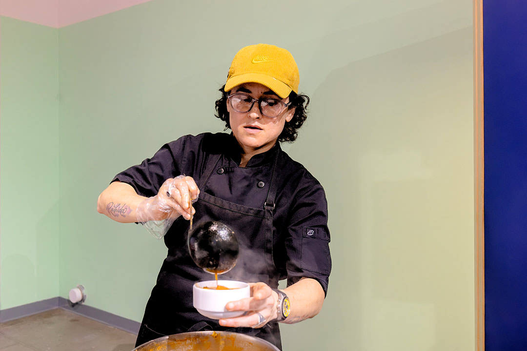 Elvira, wearing a black chef’s uniform, pours steaming birria stew from a ladle into a white bowl.