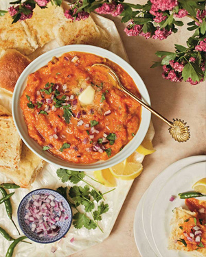 Red bhaji in a white bowl with ingredients and pav surrounding it, and pink flowers to one side.