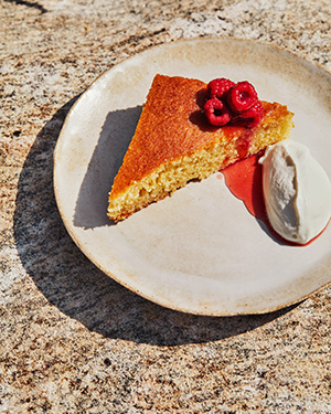 A cake on a beige plate, garnished with a handful of raspberries on top and yogurt whipped cream to the side.